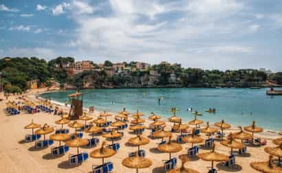 Strand von Porto Cristo auf Mallorca mit Wasser, Strandstühlen und Stadt im Hintergrund