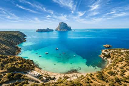 Traumhafter Blick auf die Bucht von Cala d’Hort auf Ibiza mit türkisblauem Meer, felsiger Küstenlandschaft und der beeindruckenden Felsinsel Es Vedrà im Hintergrund.