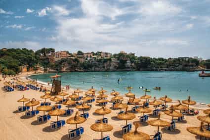 Strand von Porto Cristo auf Mallorca mit Wasser, Strandstühlen und Stadt im Hintergrund