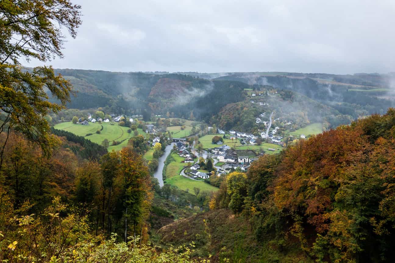 Panoramablick über das idyllische Dorf Ouren mit seinen historischen Gebäuden, umgeben von grünen Wäldern und sanften Hügeln unter blauem Himmel