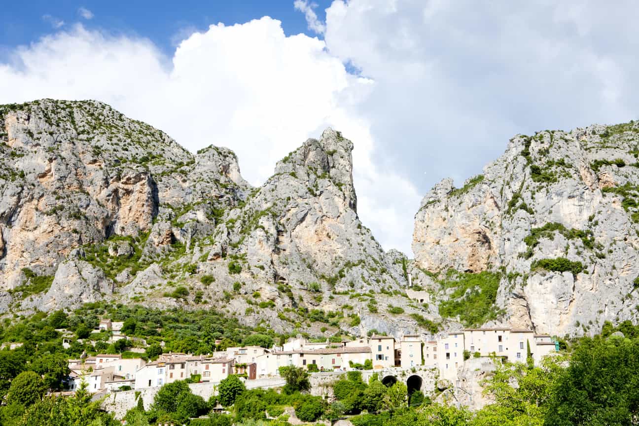 Blick von unten auf Moustiers-Sainte-Marie mit Dorf im Vordergrund und Felsen im Hintergrund