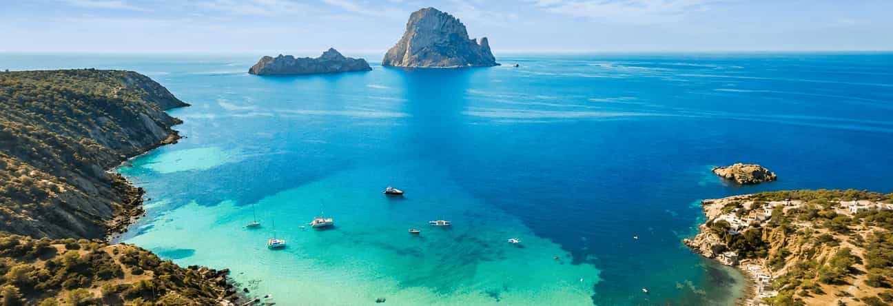 Traumhafter Blick auf die Bucht von Cala d’Hort auf Ibiza mit türkisblauem Meer, felsiger Küstenlandschaft und der beeindruckenden Felsinsel Es Vedrà im Hintergrund.